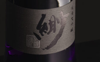日本酒『緲（びょう）』のボトルラベル。筆文字で力強くデザインされた『緲』の文字が黒い和紙風ラベルに大きく描かれ、右側に『純米大吟醸』の文字が添えられている。背景はシックな黒、ボトルは紫がかったガラス。